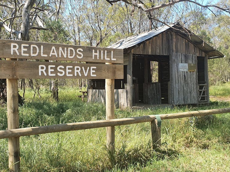 A photo of a wooden sign reading Redlands Hill Reserve. A shearing shed is behind it.