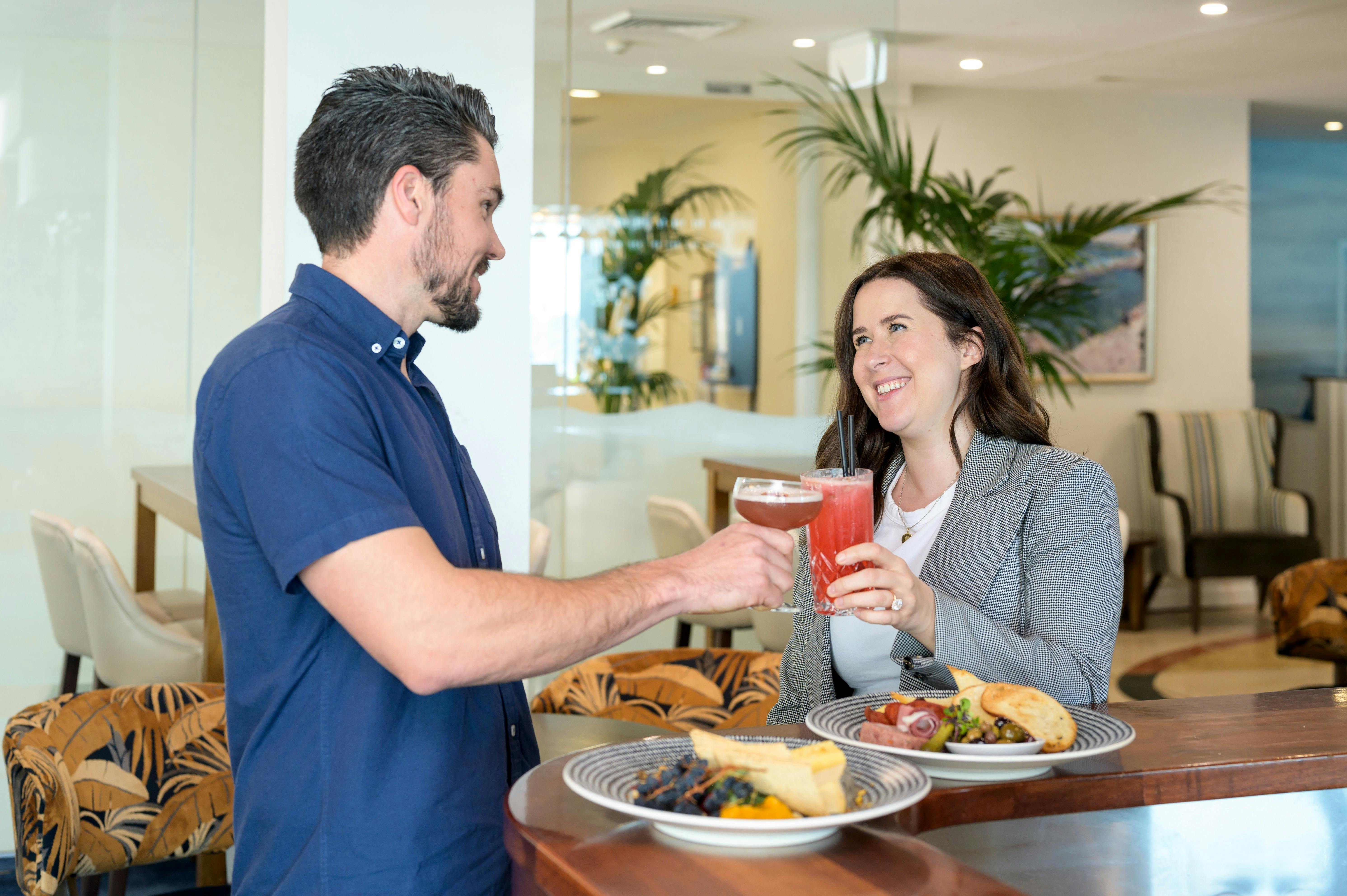 Two people enjoying cocktails and bar food standing at NOAH'S Bar. Bar furniture and palm trees