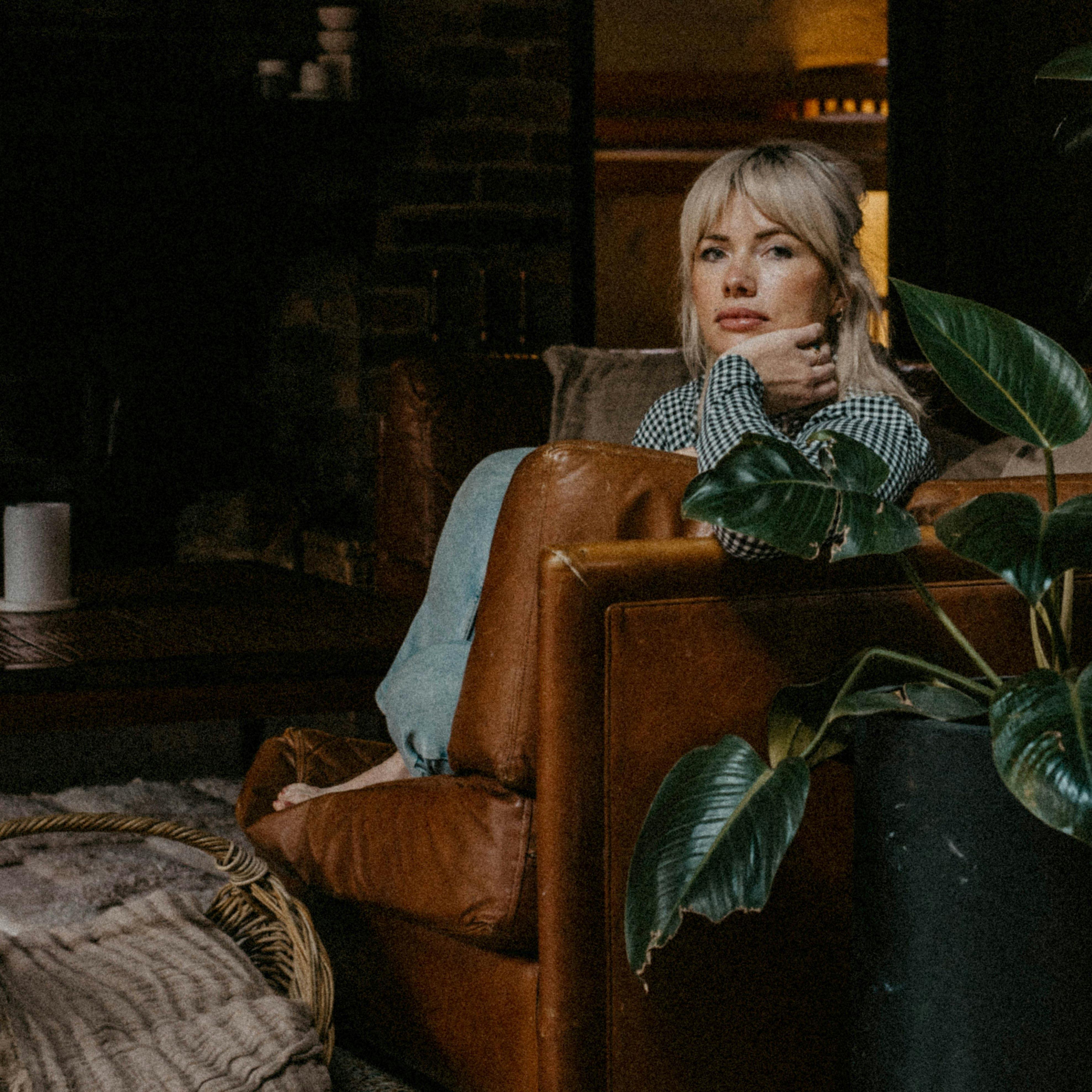 Clementine sits on a leather couch beside a potted plant in a cosy dimly lit room.
