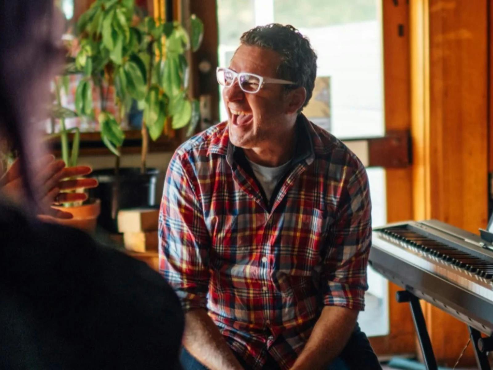 Man in plaid shirt and white glasses laughing, sitting near a keyboard and plants.