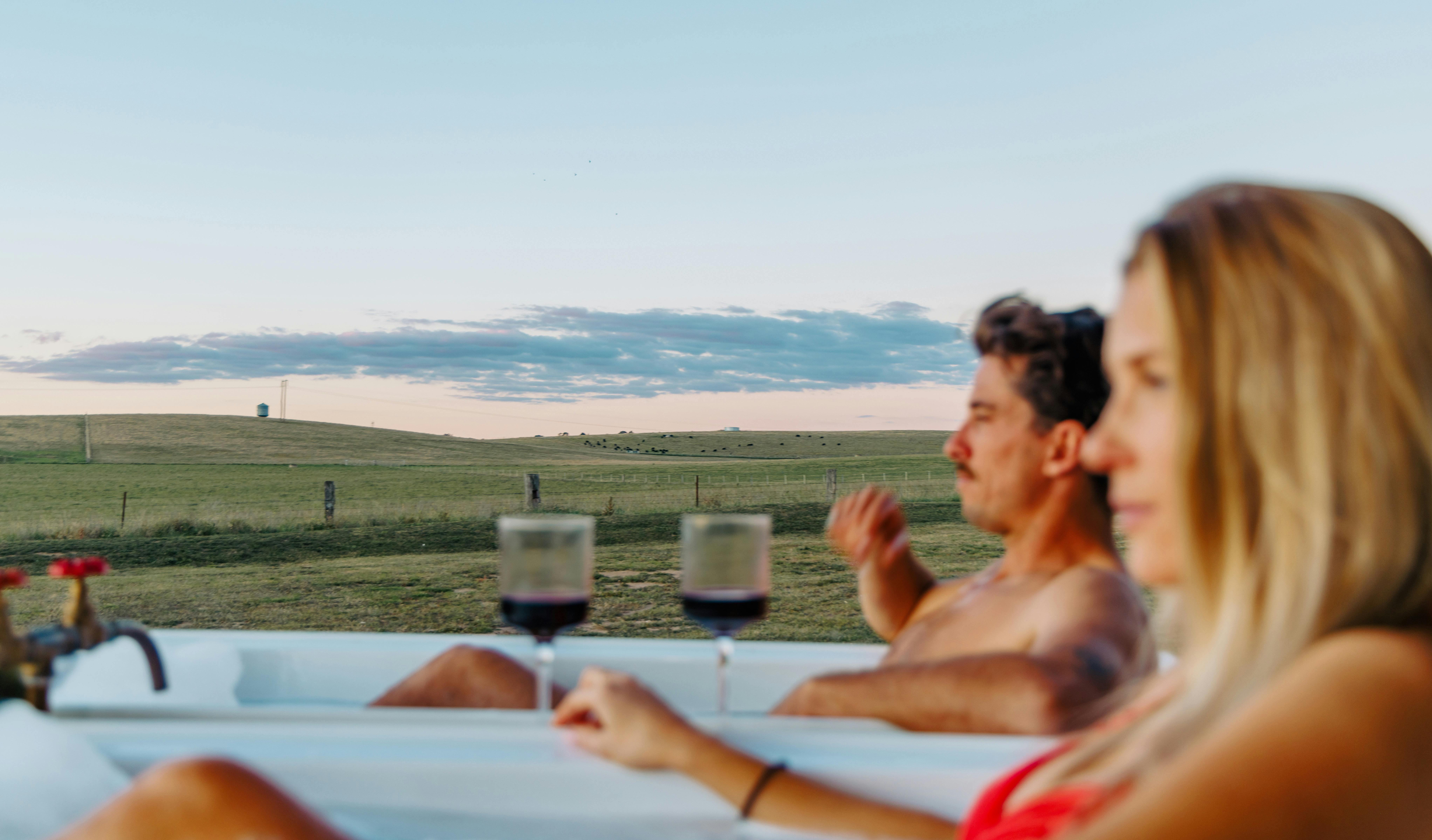 A male and female in foreground enjoying wine in bath, paddock views in background.