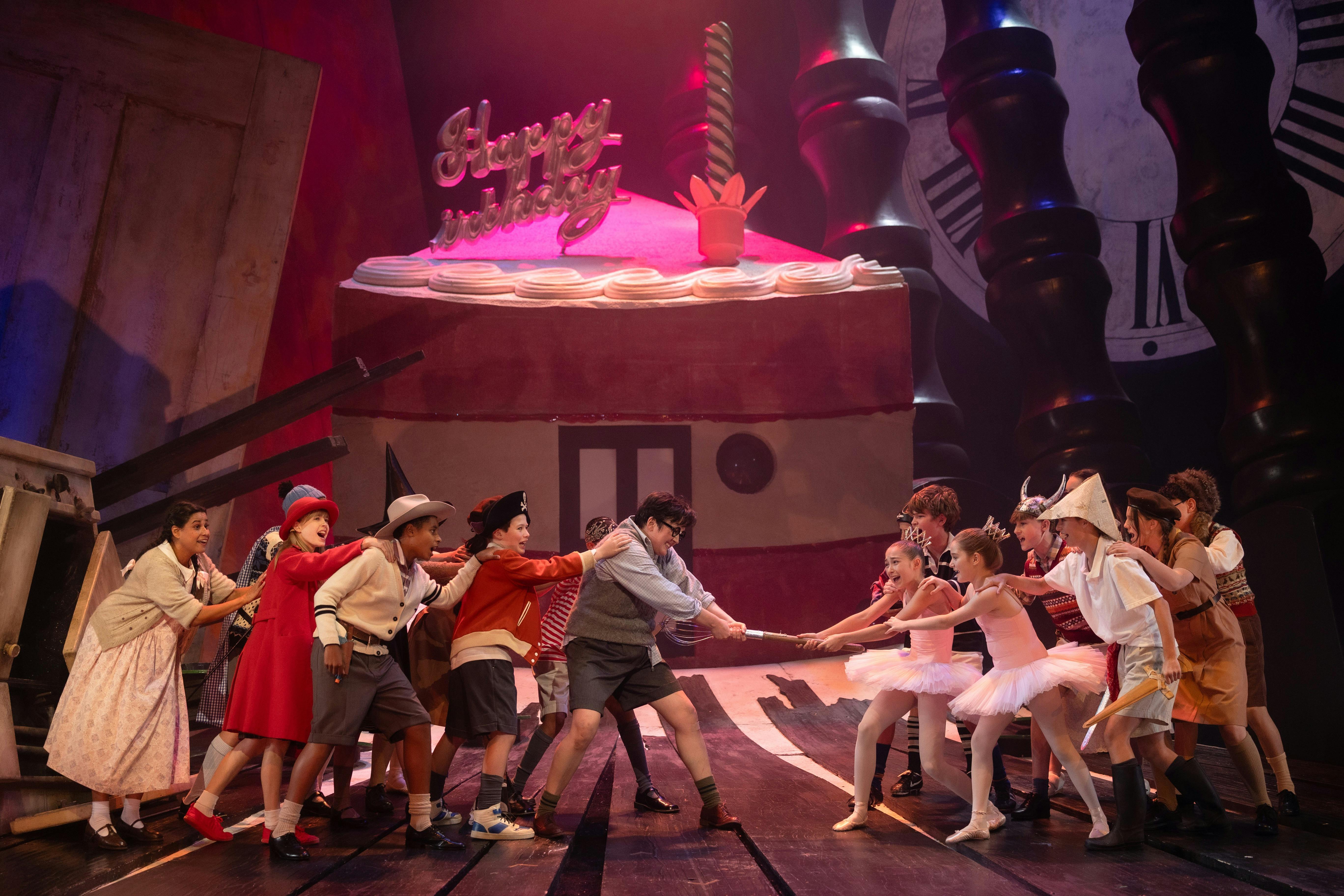 In front of a towering birthday cake, a group of children are playing tug of war with a giant whisk