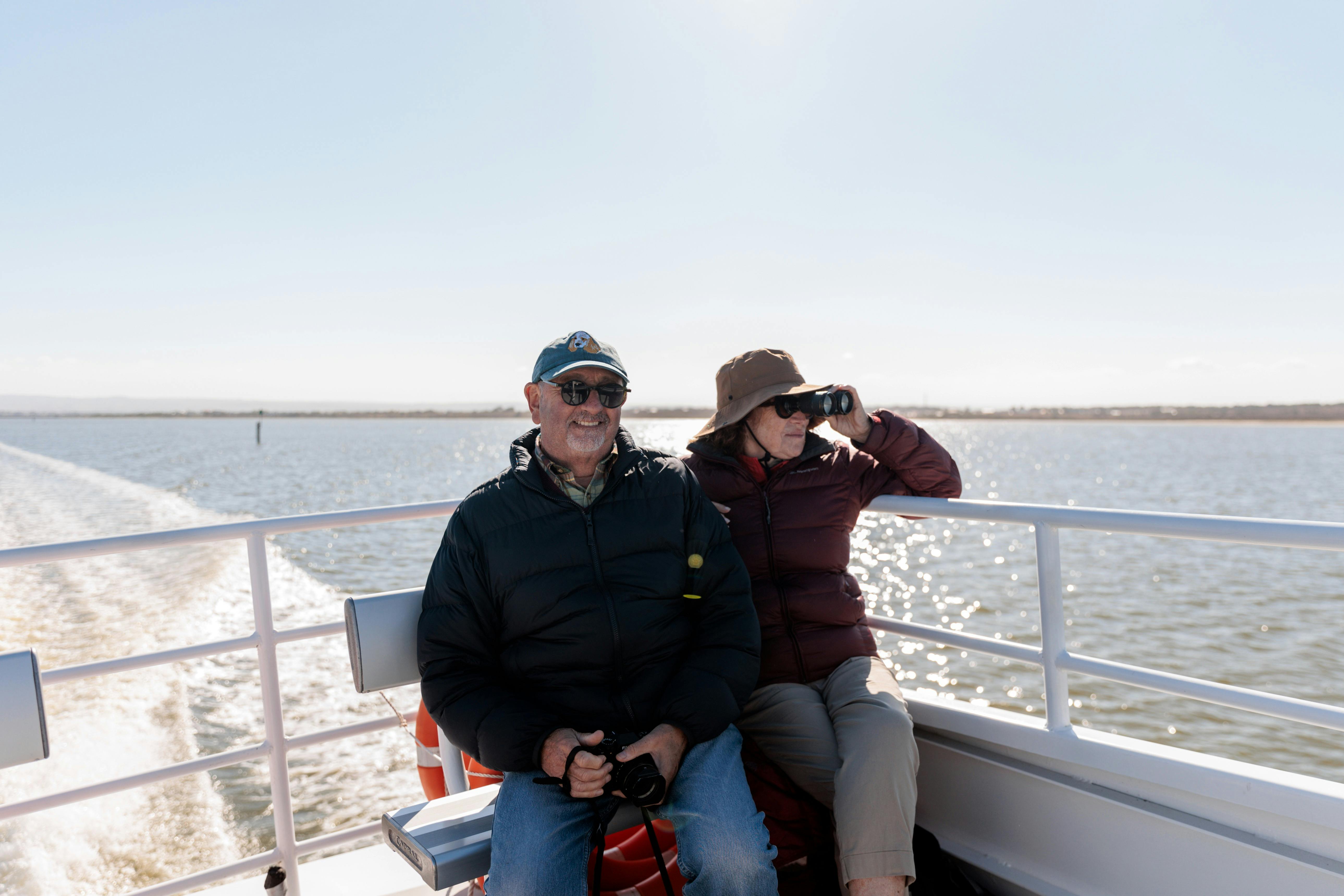An older couple enjoys a boat ride, the man smiling with binoculars, and the woman using binoculars.