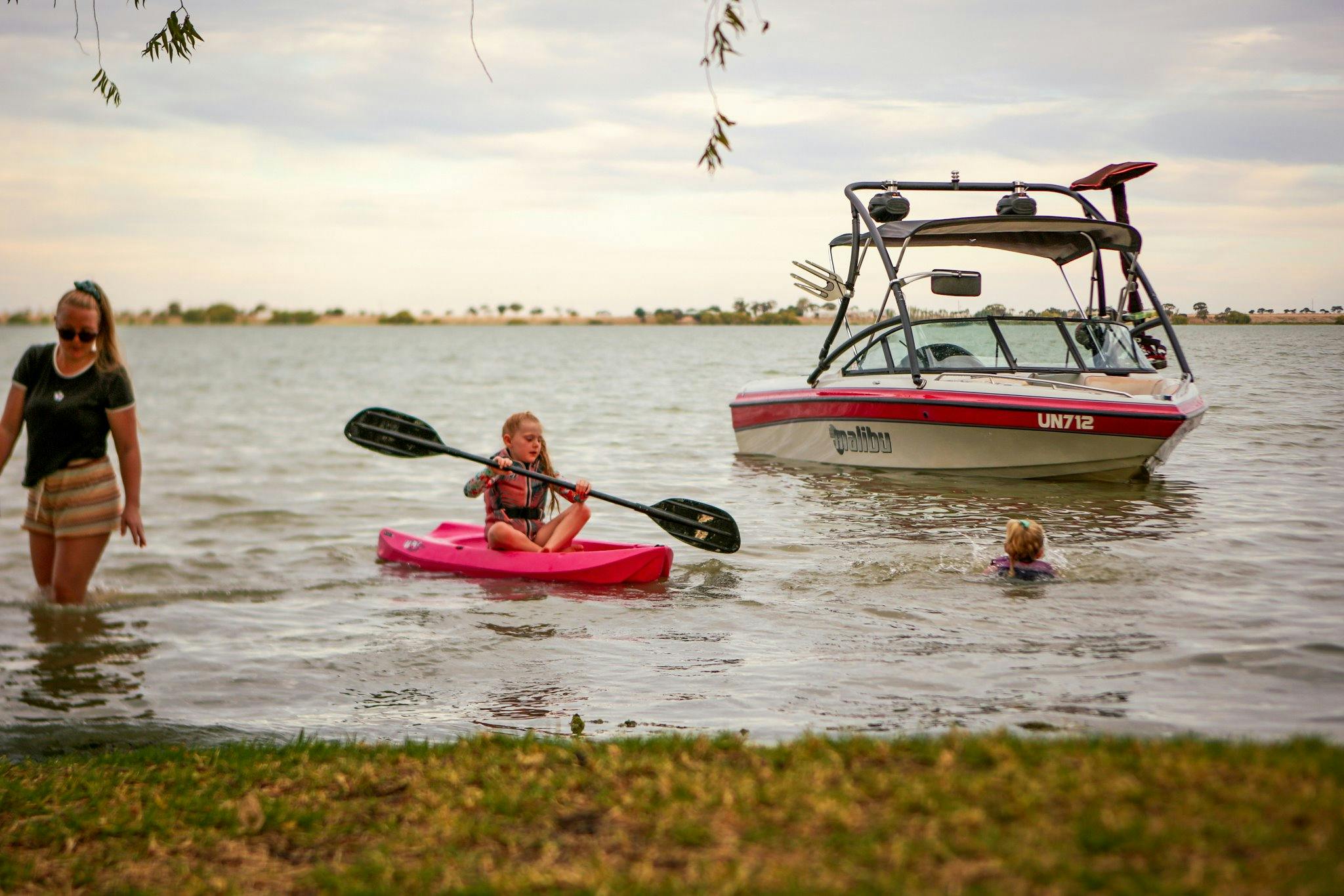 Children swimming and kayaking in shallow water with a Malibu speedboat in the background