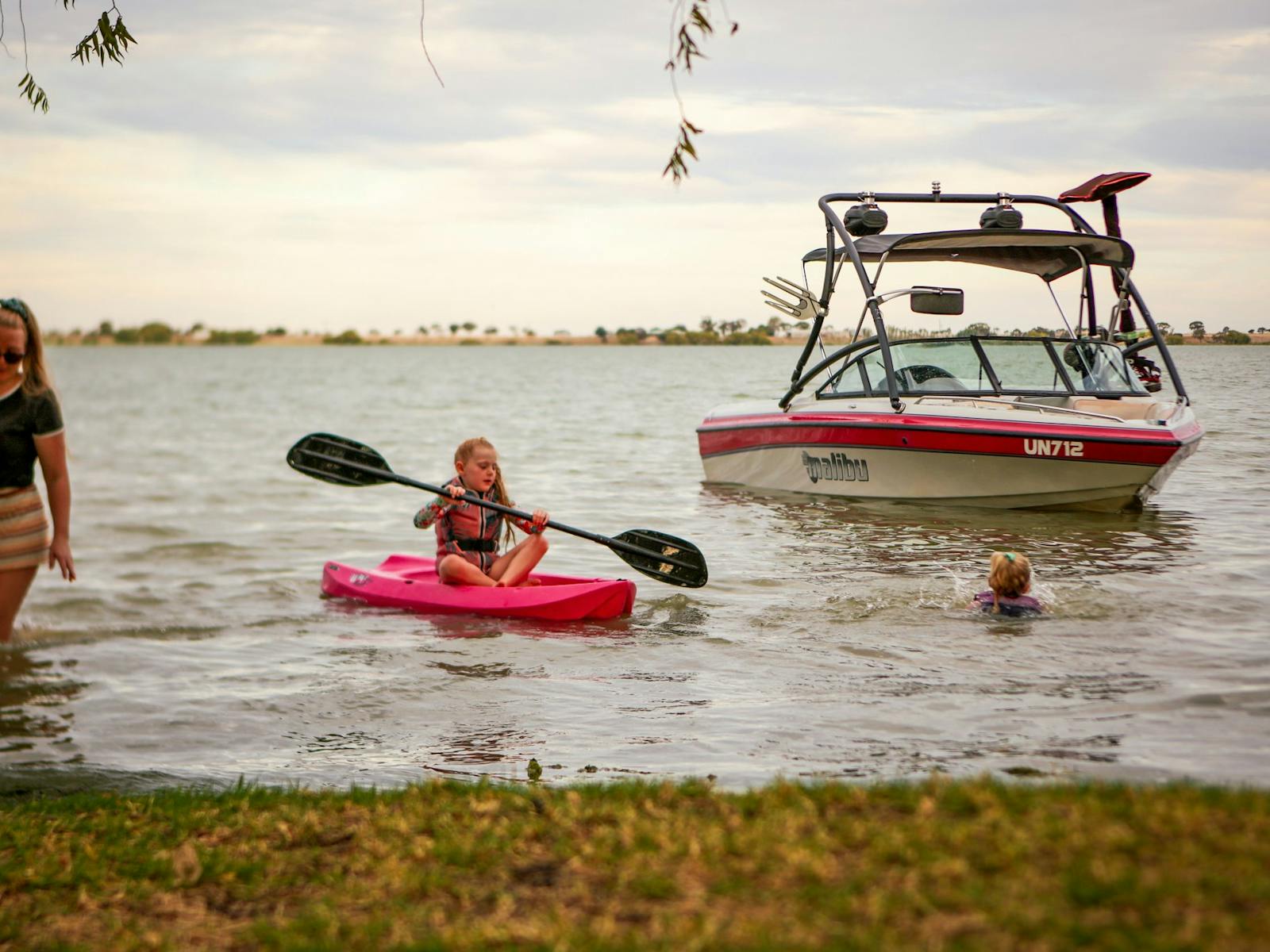 Children swimming and kayaking in shallow water with a Malibu speedboat in the background