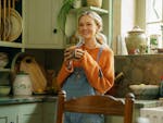 A young woman holds a cup of tea smiling, in a kitchen.