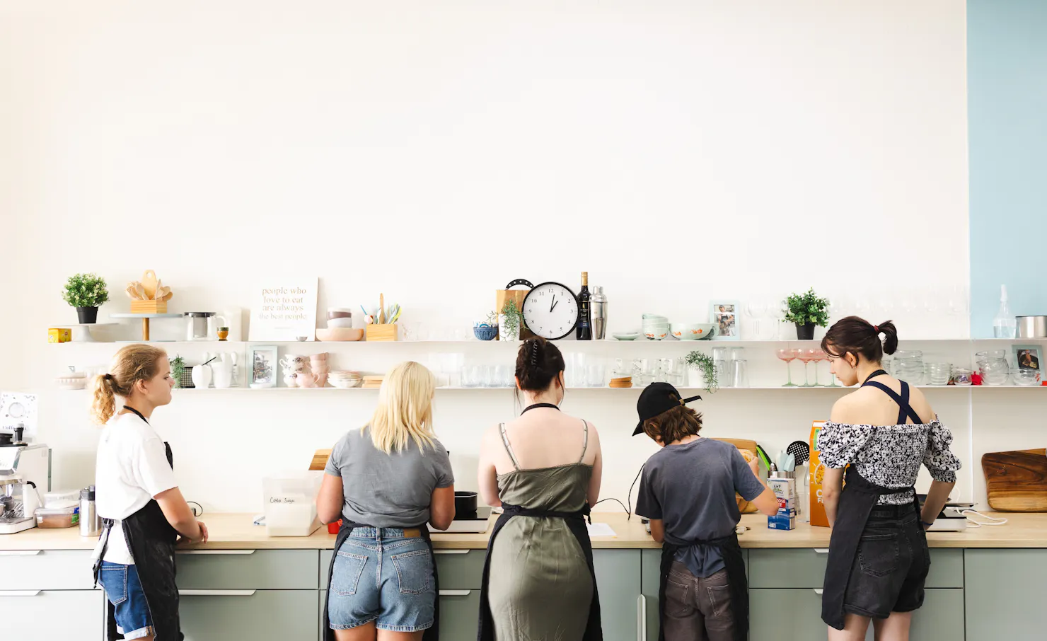 five people standing at a kitchen bench, cooking.