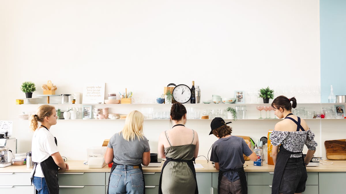 five people standing at a kitchen bench, cooking.