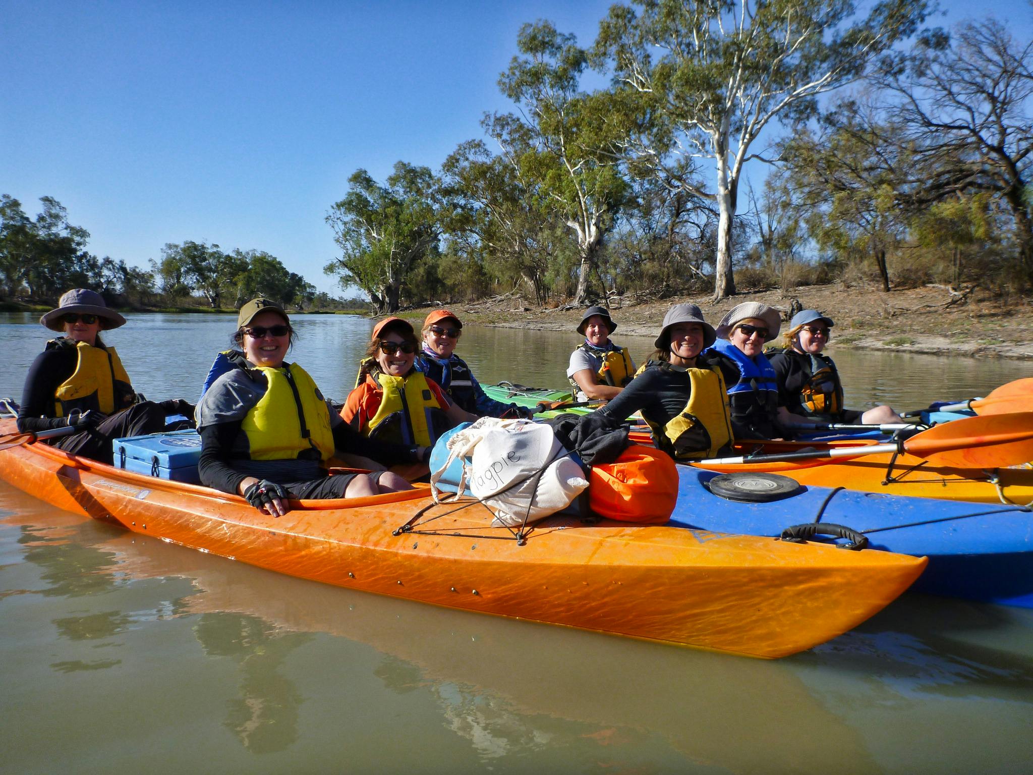 A group of smiling ladies wearing life jackets, in double kayaks, loaded with camping gear.