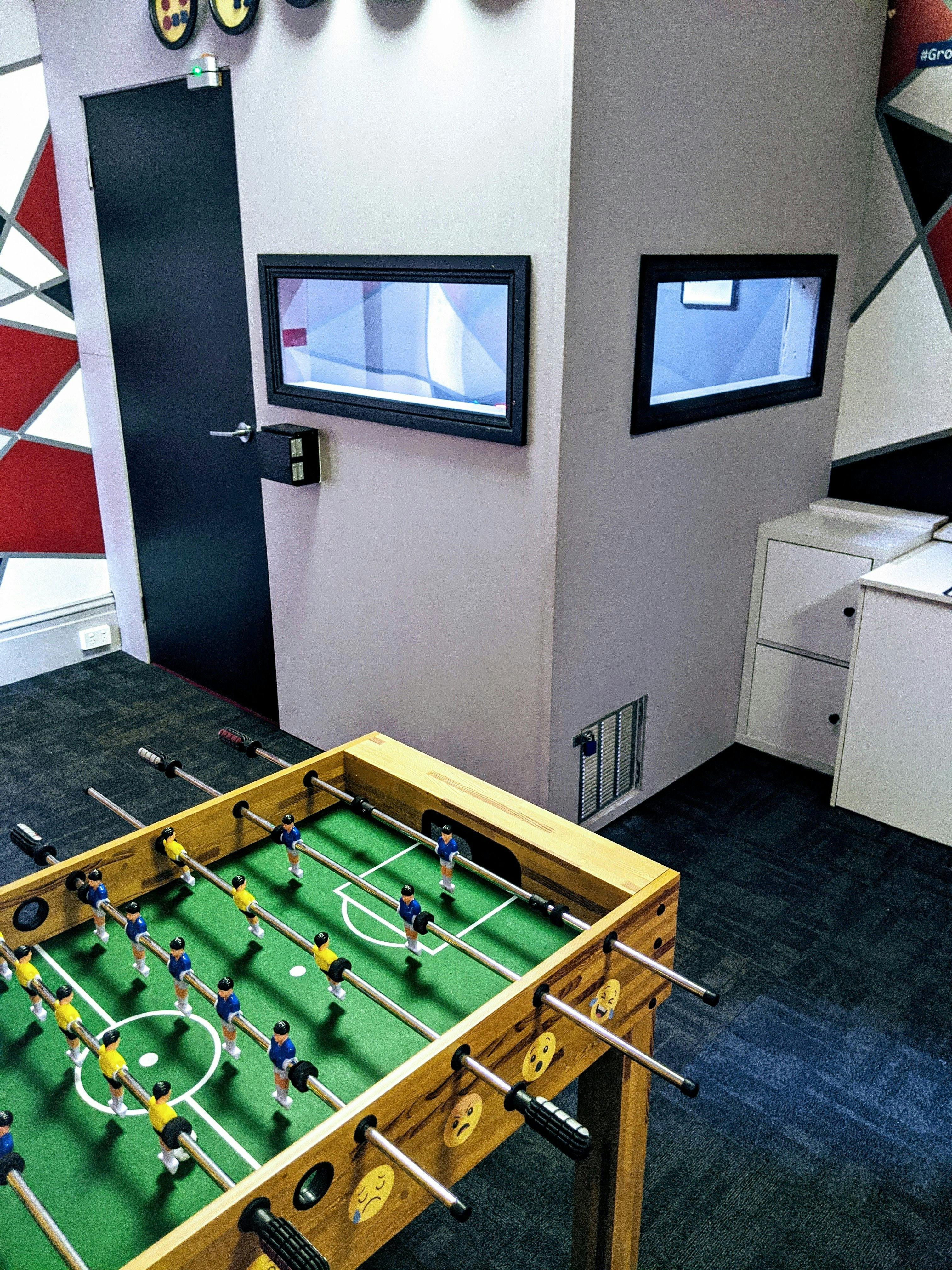 Side view of a foosball table with a desk and small grey room in the background, with two windows