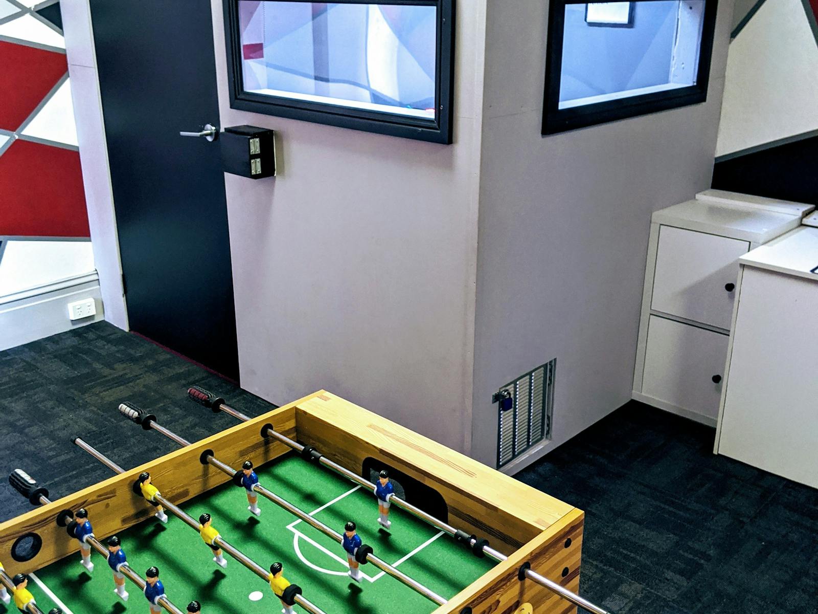Side view of a foosball table with a desk and small grey room in the background, with two windows