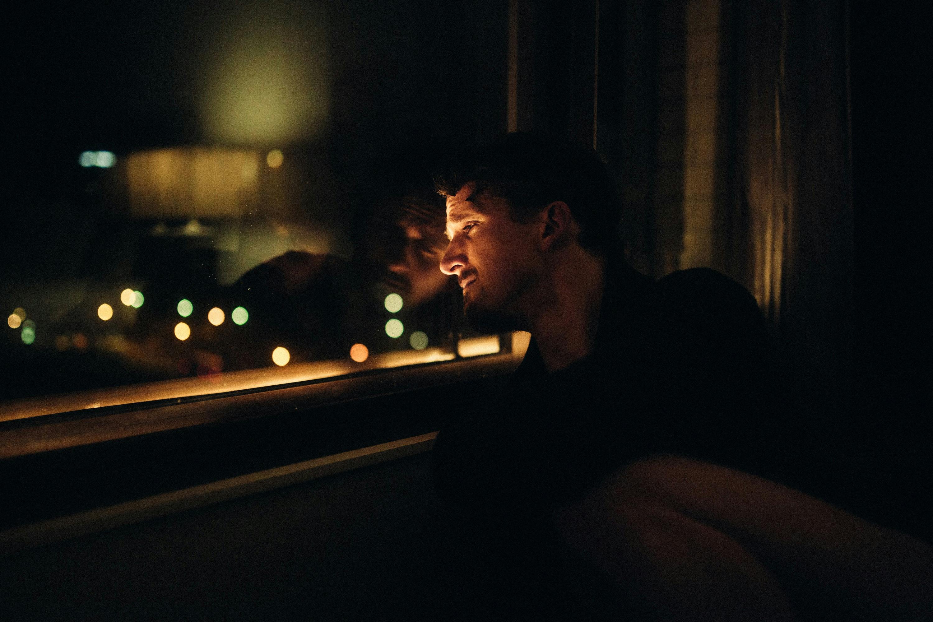A man gazes out a window at night, his reflection visible, surrounded by soft, out-of-focus lights.