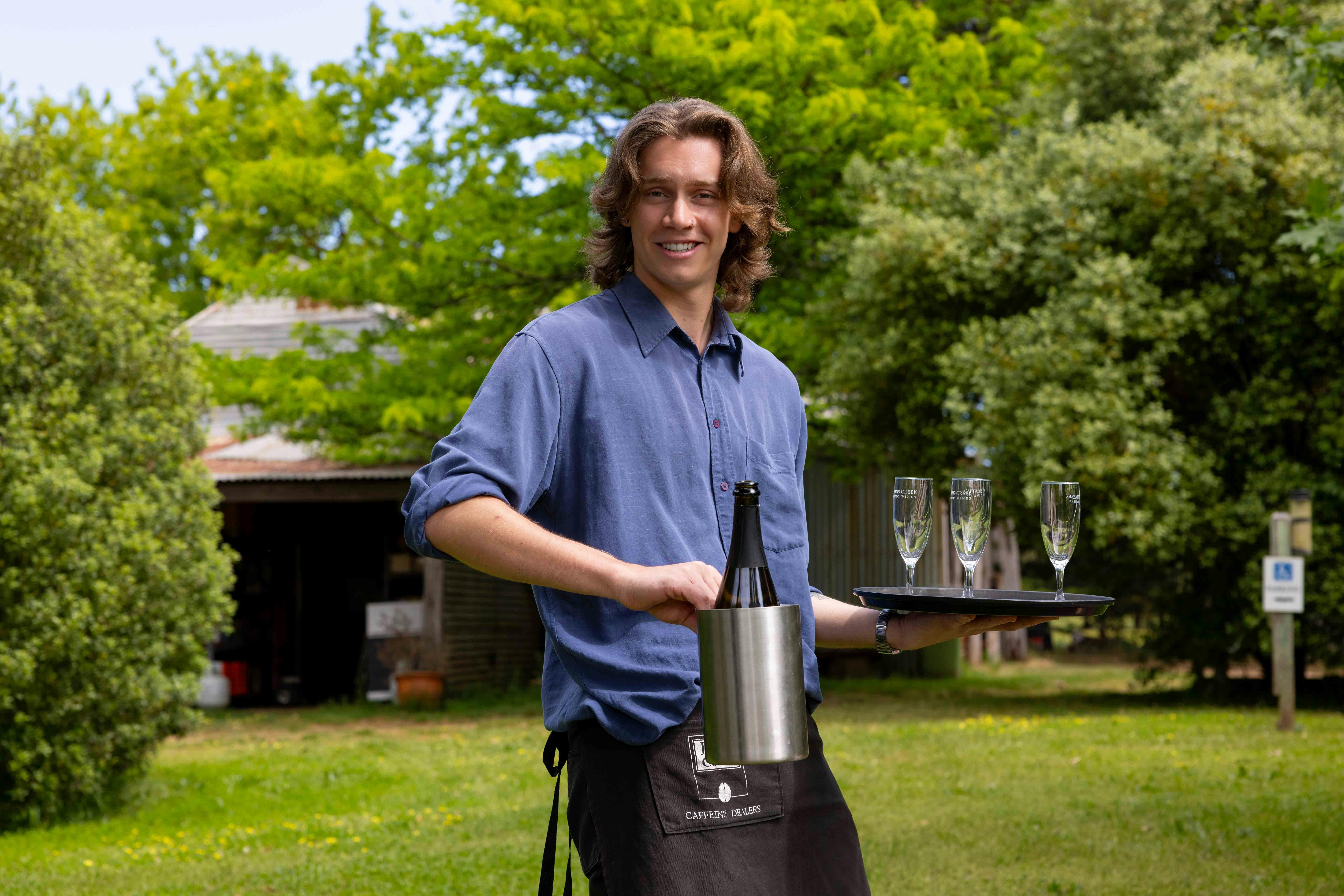 a young male carries an ice bucket with sparkling wine and wine glasses.  Grass and trees behind him