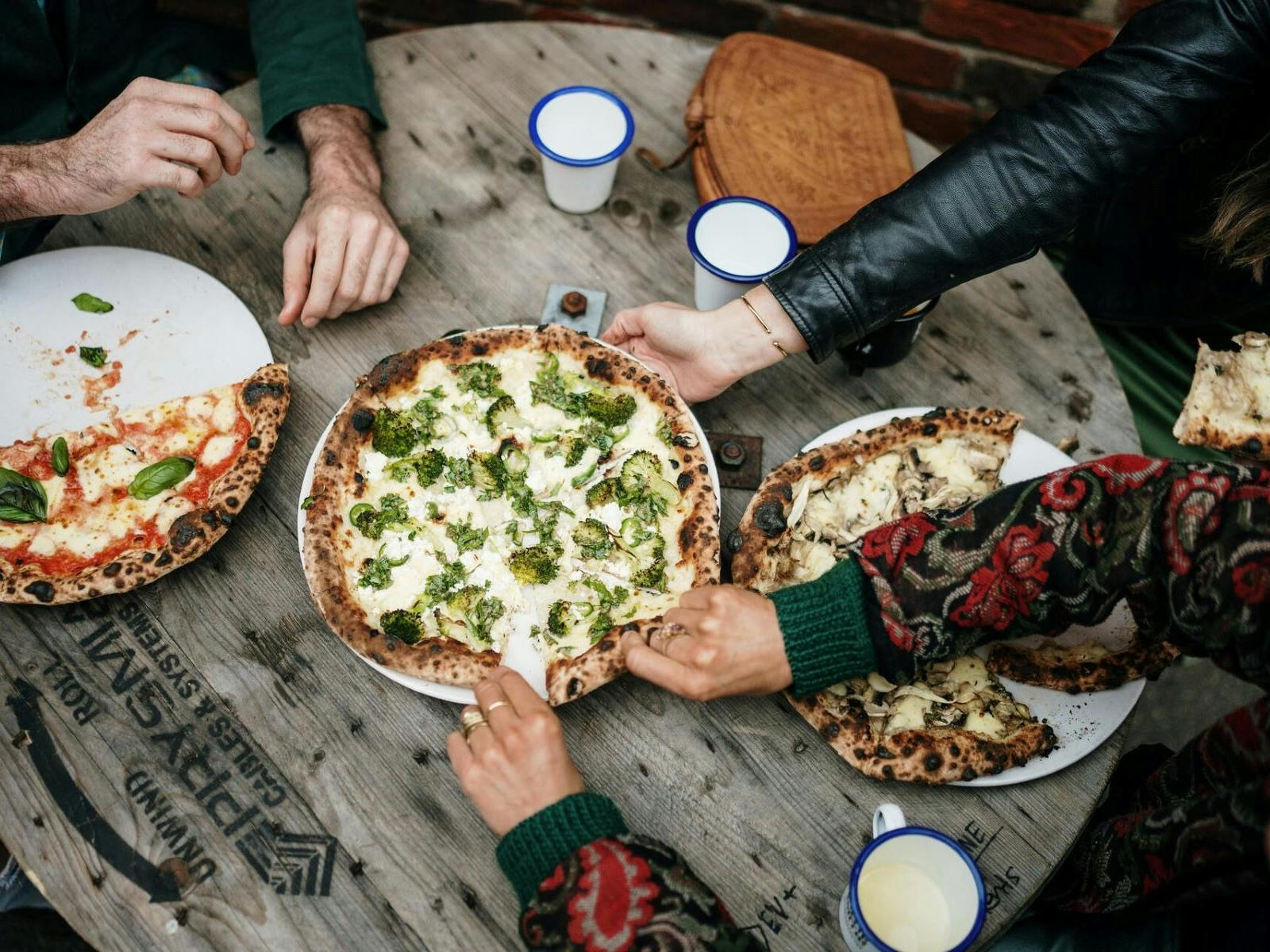 Three cheesy pizzas, one half eaten with  people reaching for pieces sitting at a round wood table