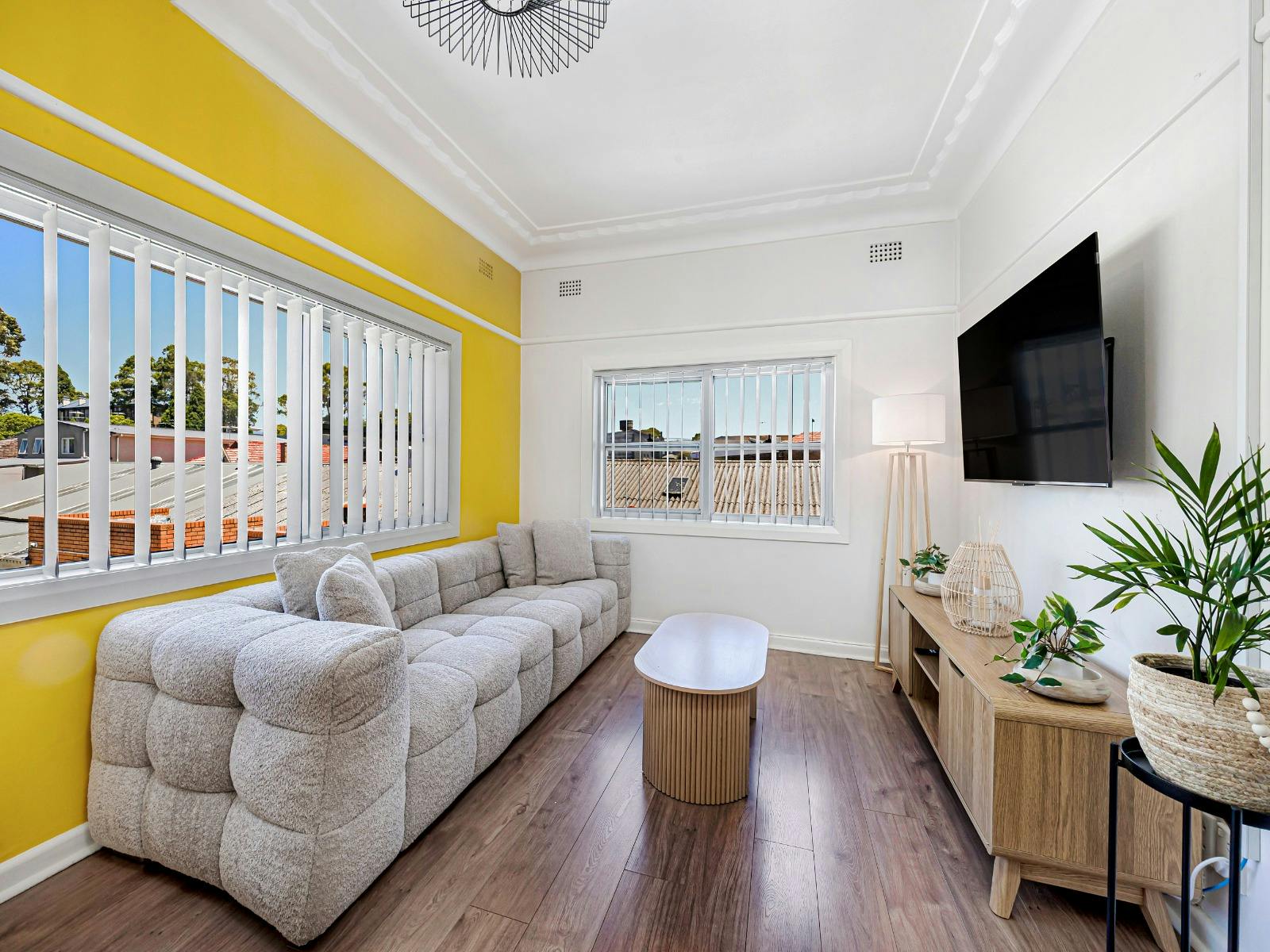 Living area with sofa, wall-mounted TV, and large windows with yellow feature wall.