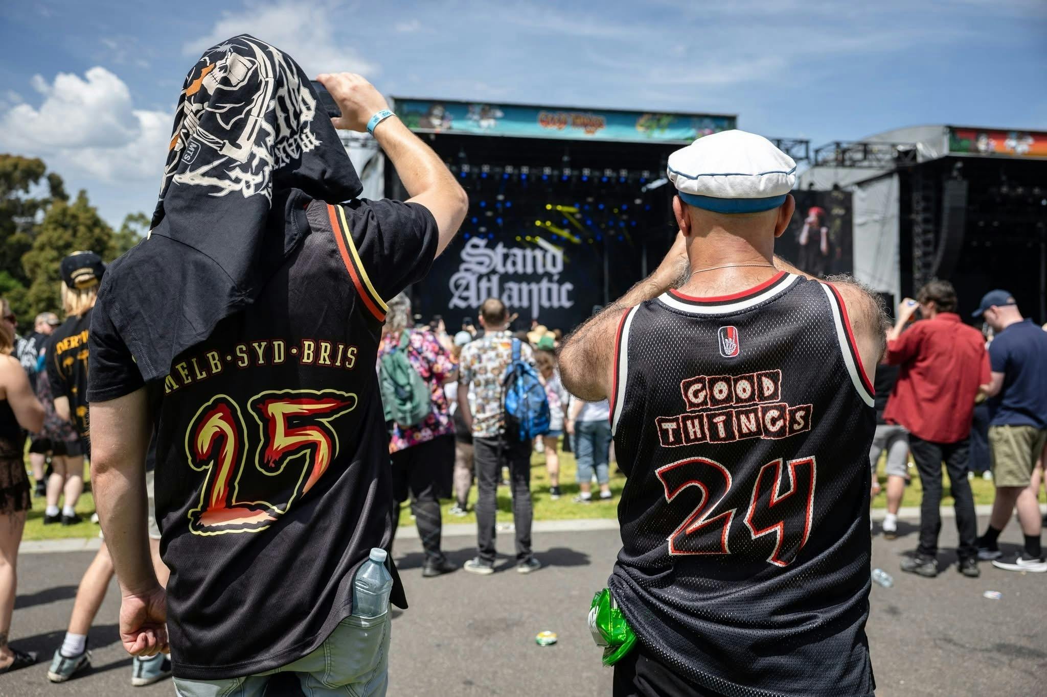 Two festival-goers in Good Things branded jerseys watching a band perform on stage under a clear blu