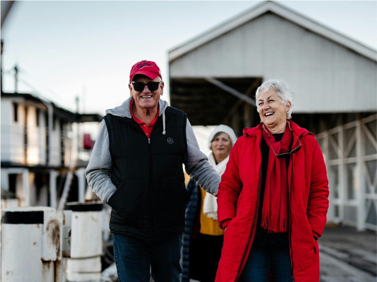 An older couple in jackets smile and walk together on a dock, with a woman in the background.
