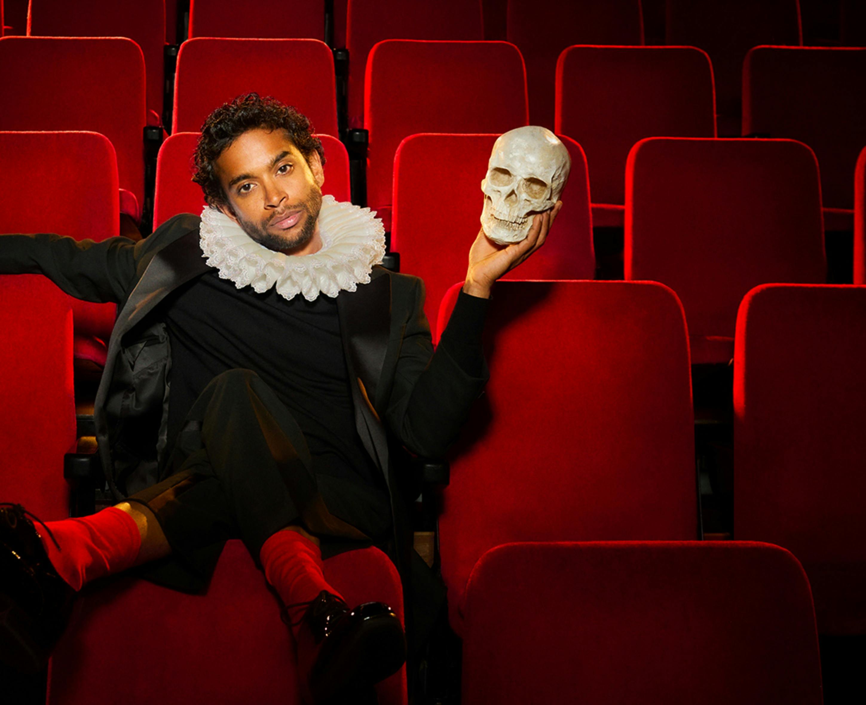 A man in a ruff holding a skull sitting in red theatre seats
