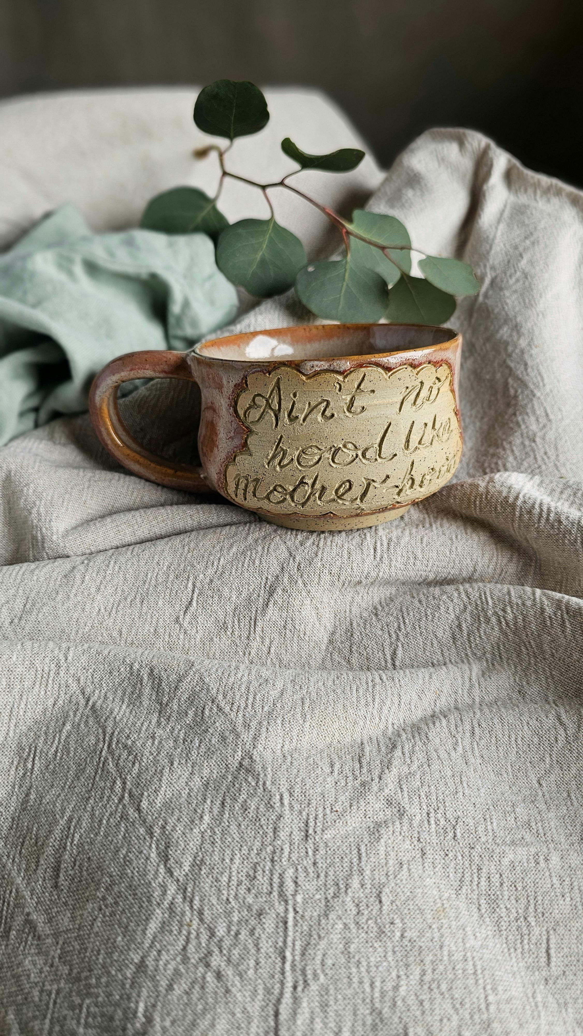 A pink cappuccino mug with a handle that says "ain't no hood like motherhood"