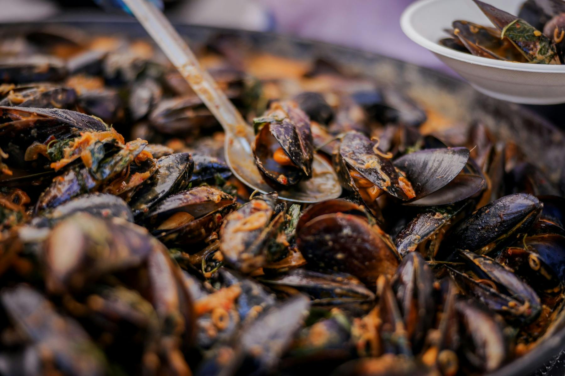 A close-up view of a dish filled with cooked mussels garnished with herbs, with a spoon scooping .