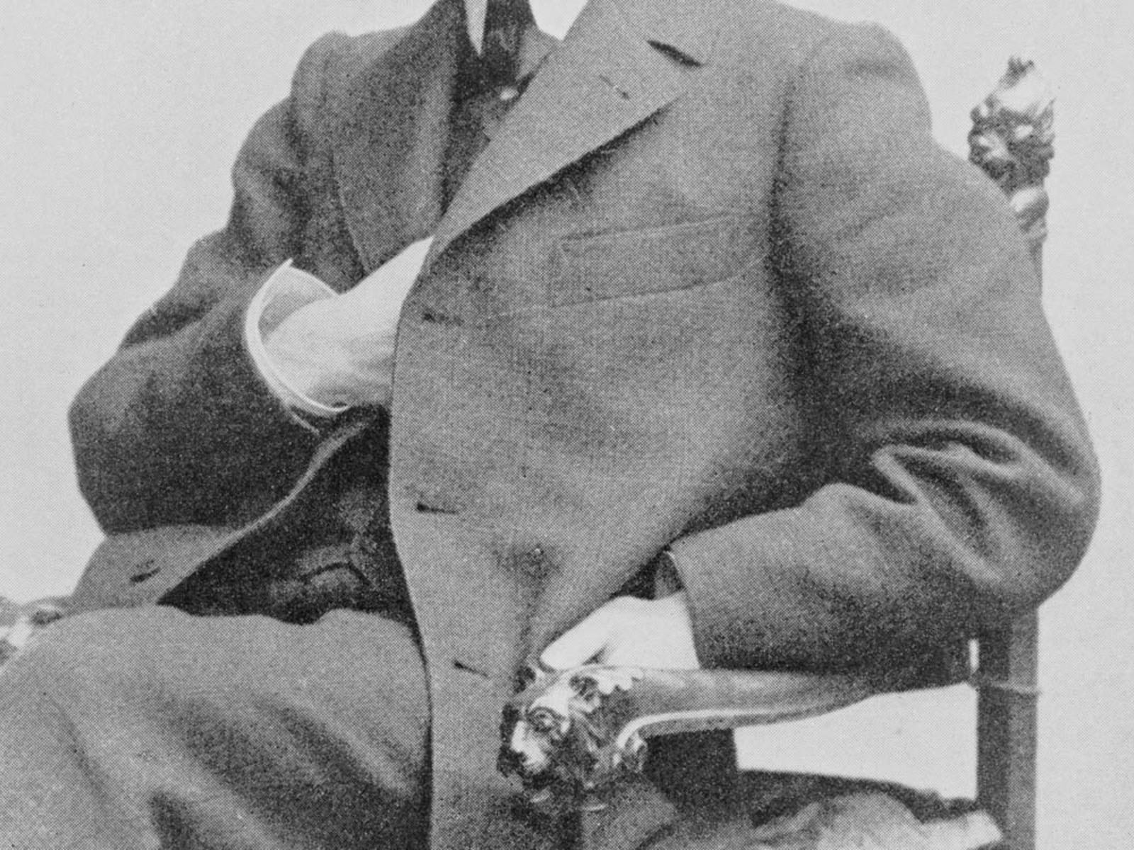 A black-and-white photograph of a man in a suit sitting in an ornate chair, with a hand tucked away
