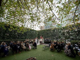 A wedding ceremony taking place in a leafy hedged courtyard with city skyline behind