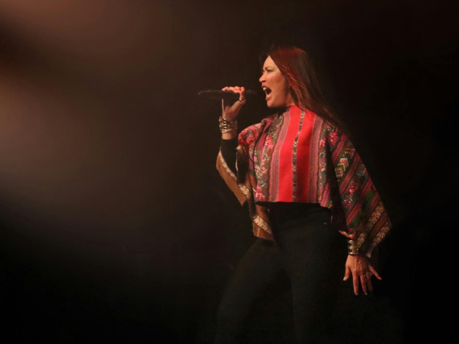 A woman in a colourful shirt and black pants stands against a brown background, singing into a mic