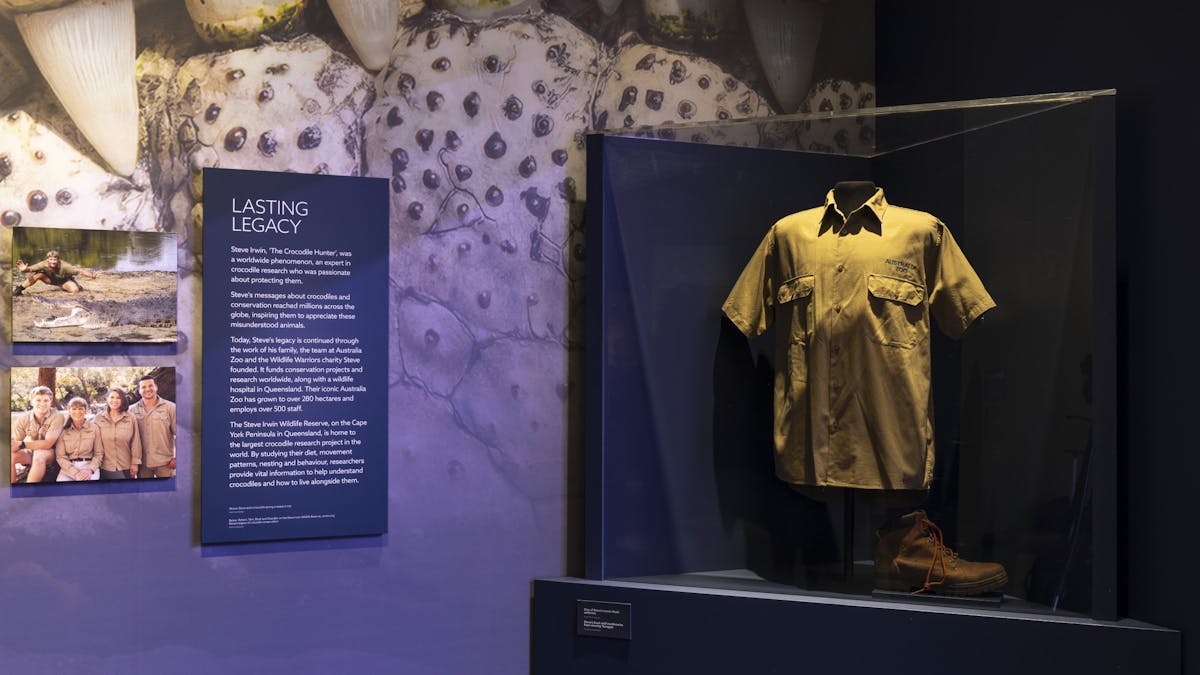 Steve Irwin's khaki uniform displayed in a museum case in front of a large crocodile graphic Steve Irwin's khaki uniform displayed in a museum case in front of a large crocodile graphic