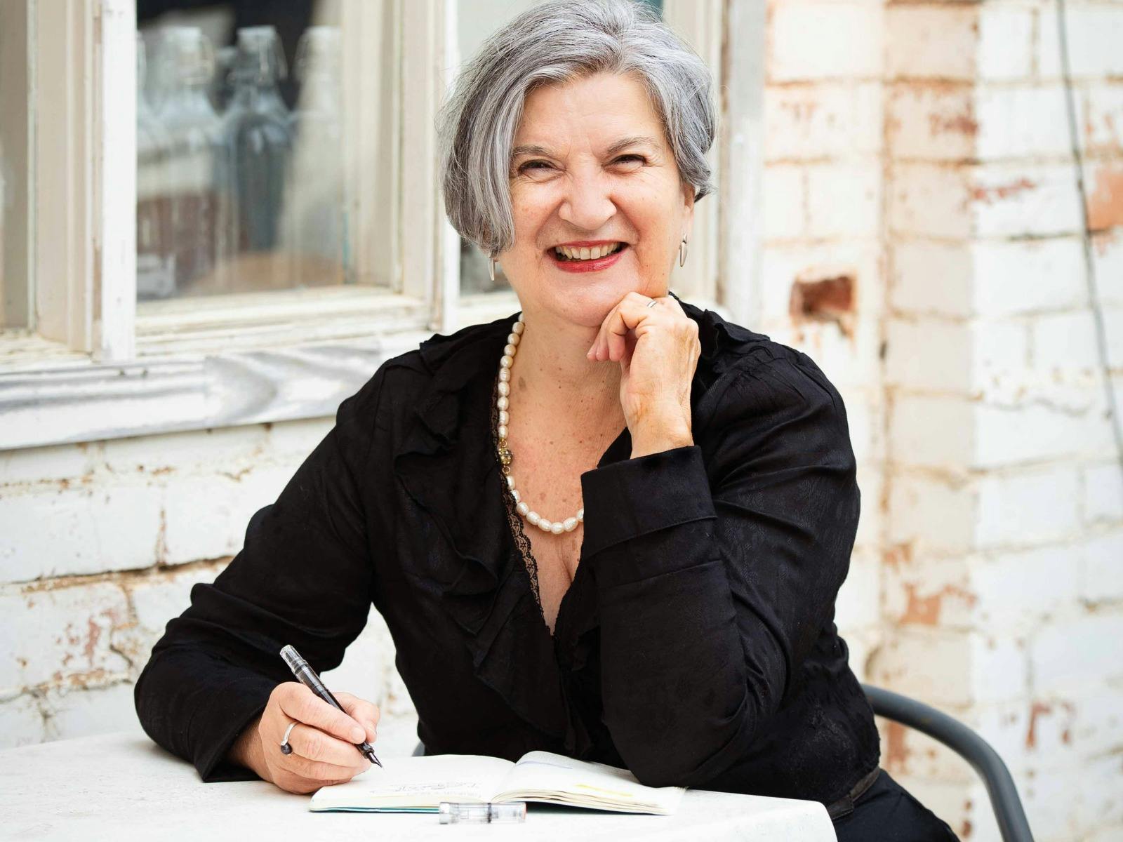A smiling woman with grey hair sits at a table outdoors, writing in a notebook.