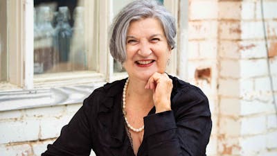 A smiling woman with grey hair sits at a table outdoors, writing in a notebook.