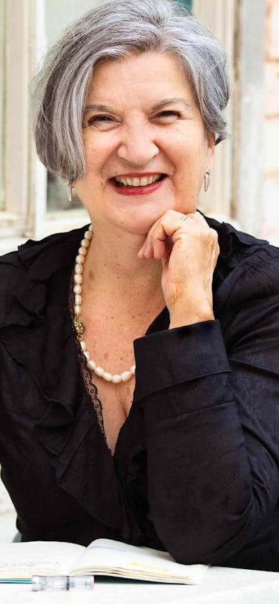 A smiling woman with grey hair sits at a table outdoors, writing in a notebook.