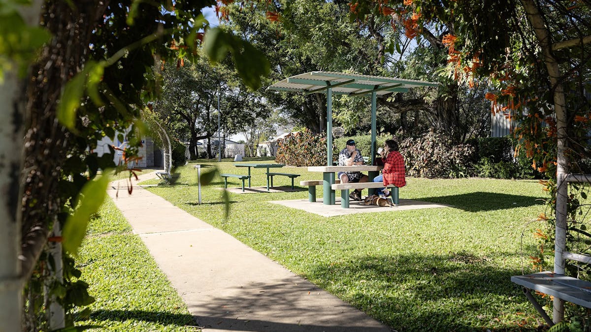 A Couple sitting on the table and chairs that has a shade over it, with footpaths,grass and trees