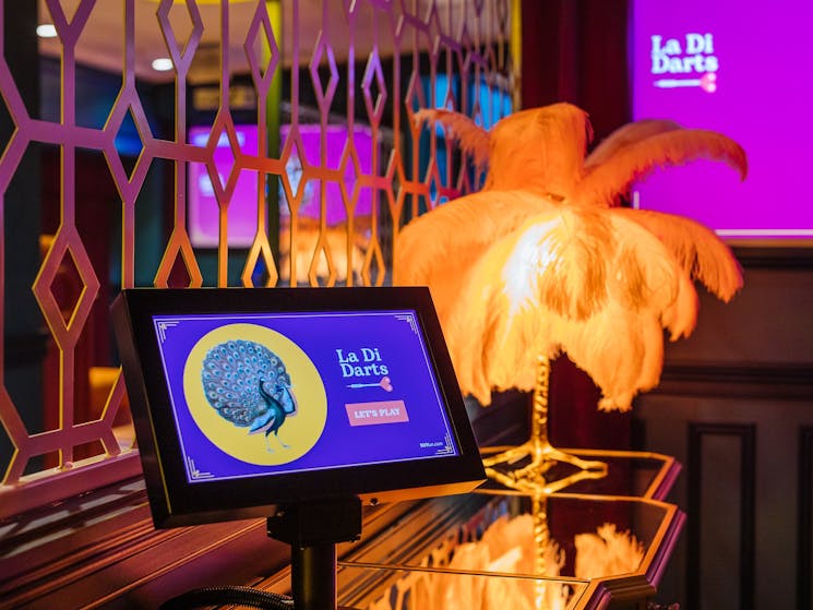 Screen displays La Di Darts logo. Decorative lamp on table.