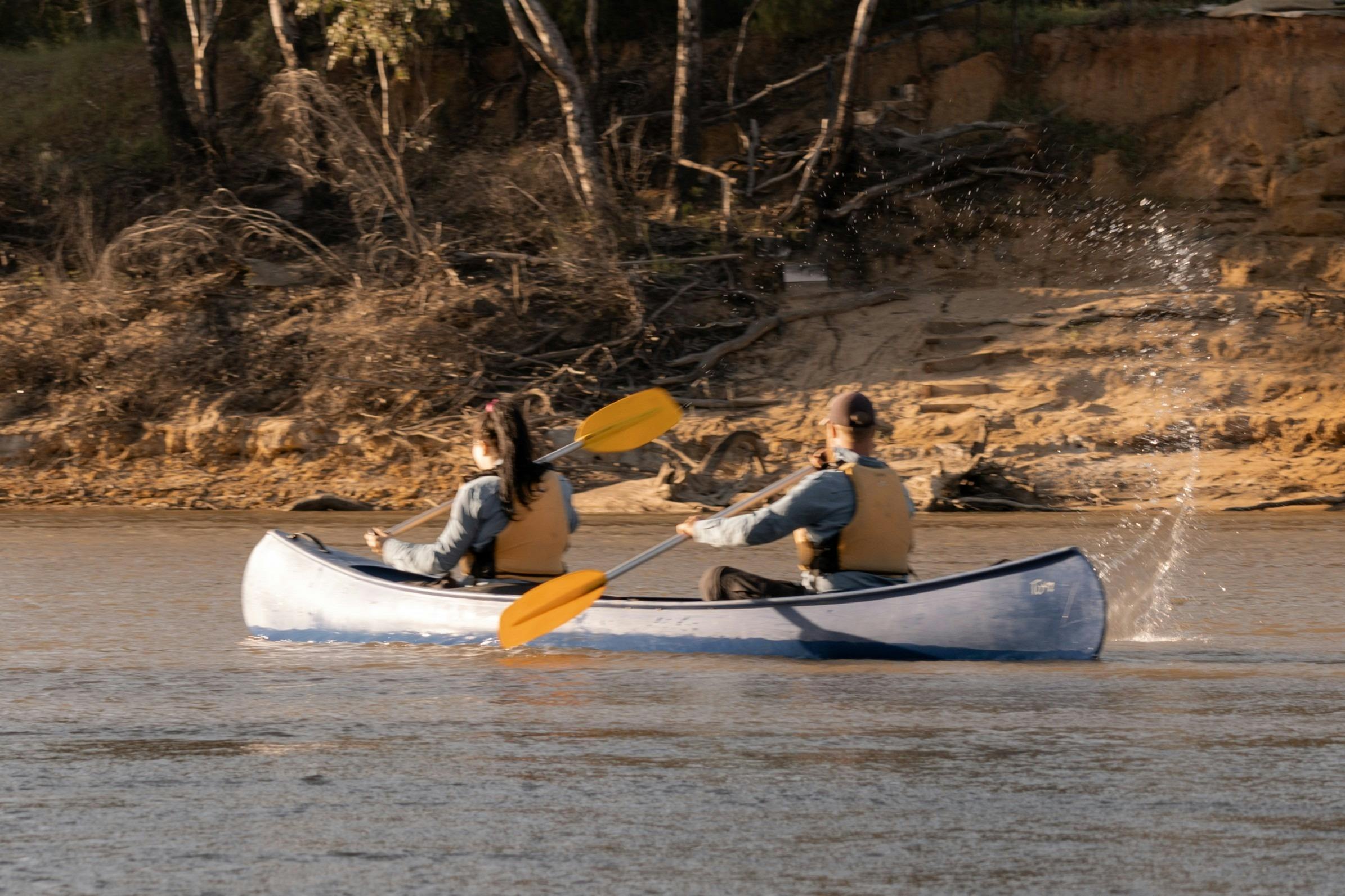 Two people wearing life jackets paddle a white canoe past a sandy riverbank and fallen trees.