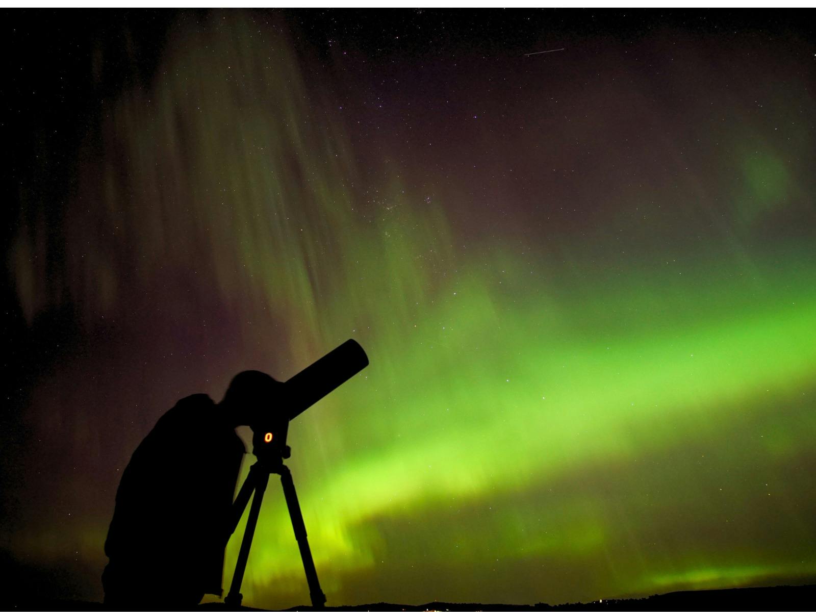 Person looking into a telescope with the aurora glowing across the night sky.