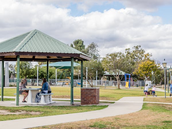 A group of women sit at a bench under a rotunda. Children ride scooters along a pathway.