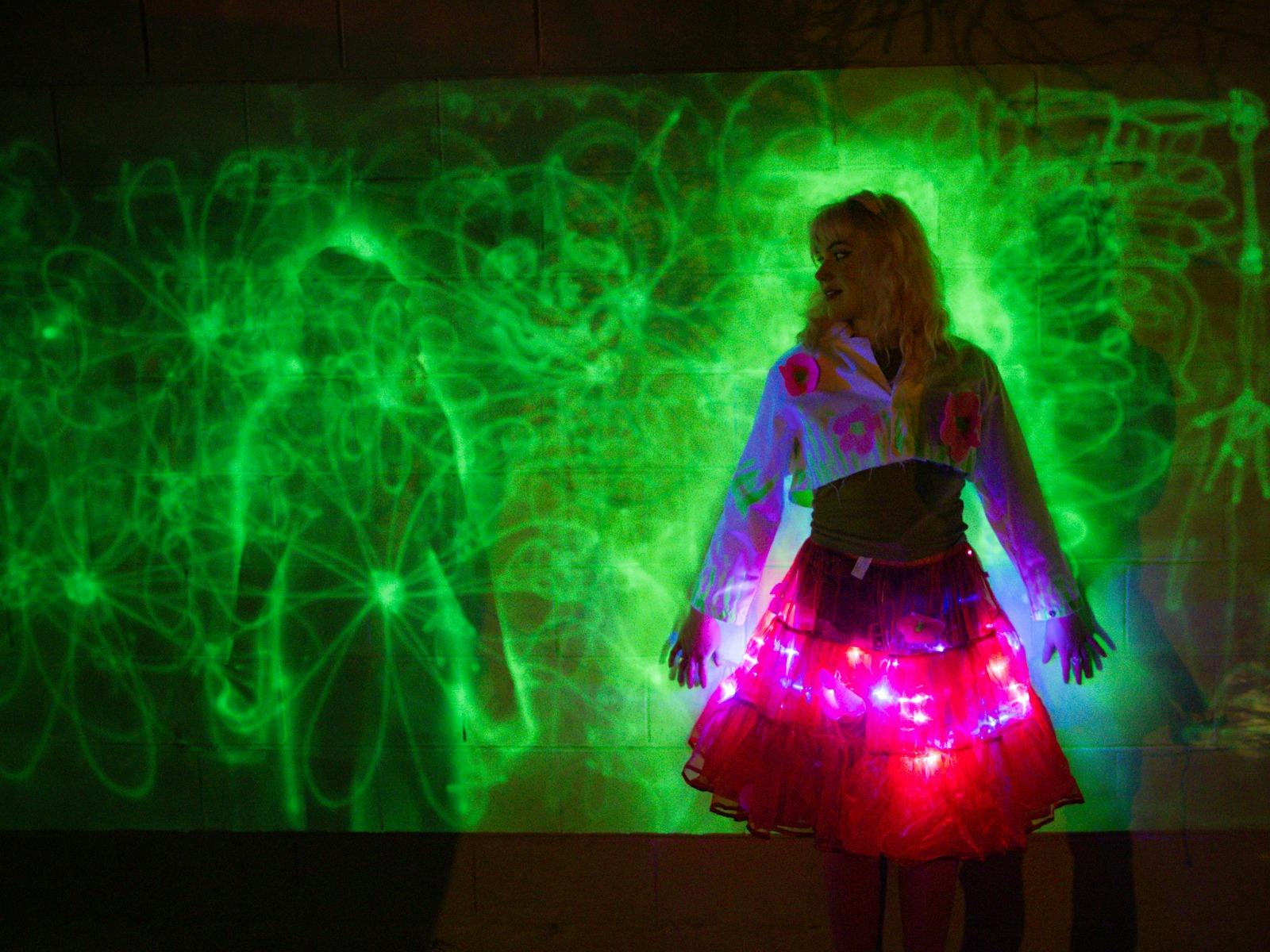 A brightly costumed woman stands against a wall with fluro-green light drawings.