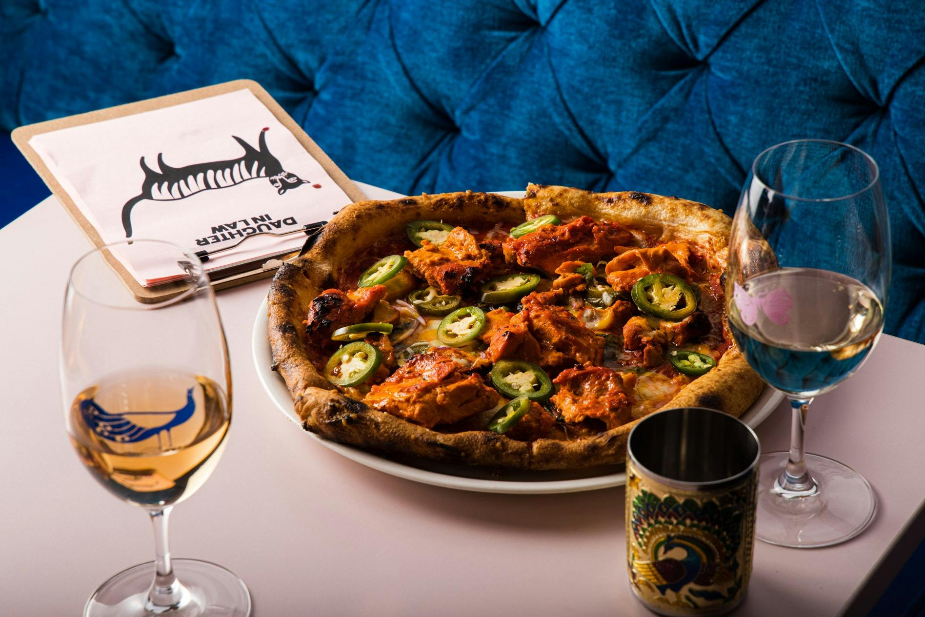 Unauthentic Tandoori chicken Naan Pizza sitting on a table with two glasses of wine and a menu