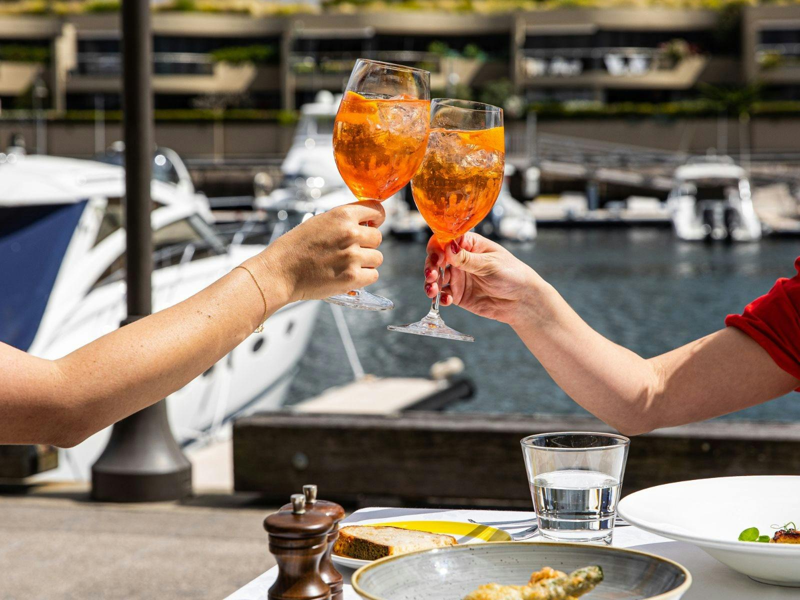 물가 항구 backgorund와 함께 환호하는 두 개의 Aperol Spritz.