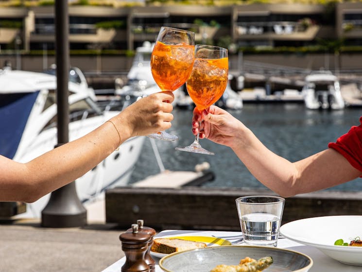 Two Aperol Spritz in a cheers' motion with a waterside harbour backgorund.