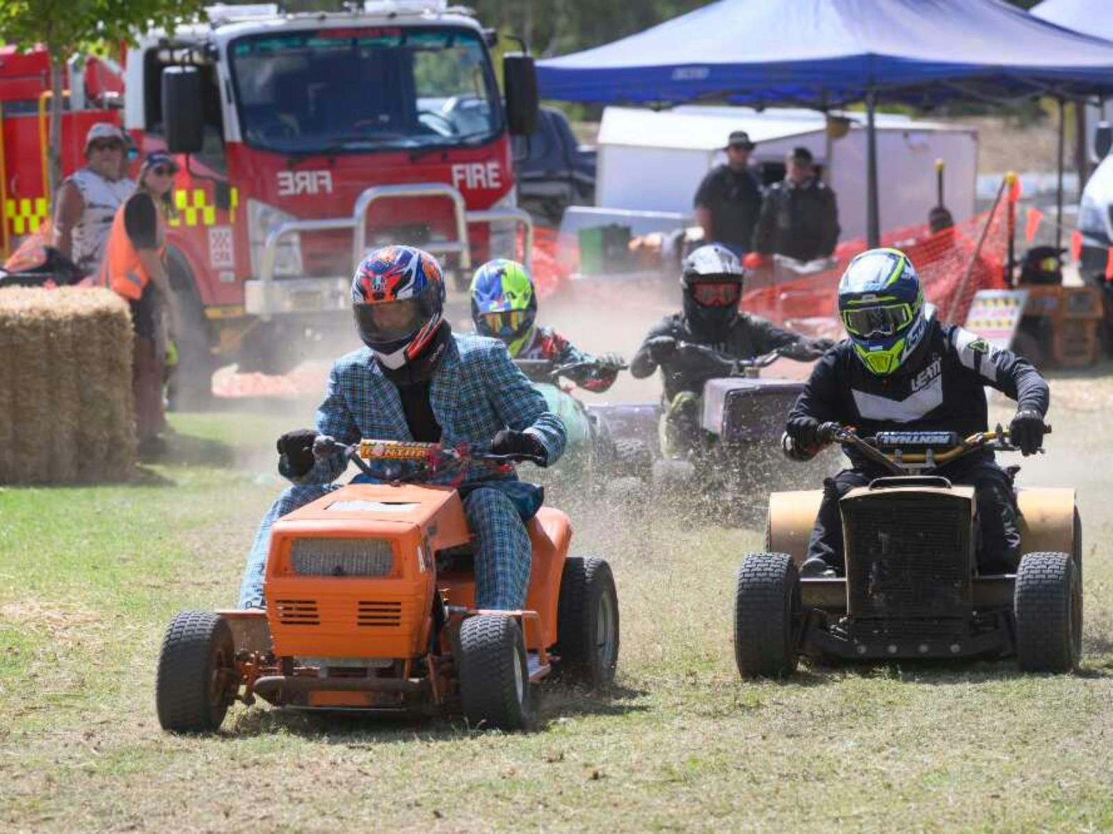Competitors race with lawnmowers on a grass track with a fire truck and spectators visible.