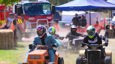 Competitors race with lawnmowers on a grass track with a fire truck and spectators visible.