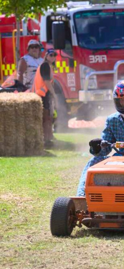 Competitors race with lawnmowers on a grass track with a fire truck and spectators visible.