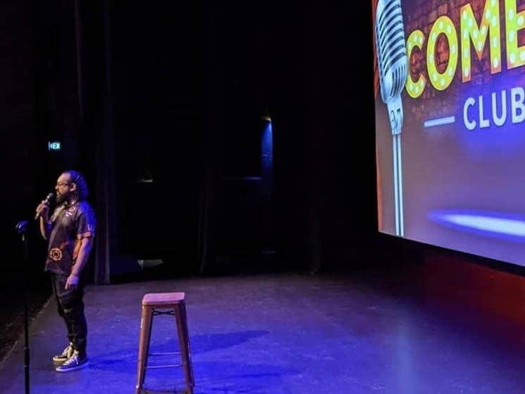 Comedian standing in front of a stool with a screen saying Casula Comedy Club at the back