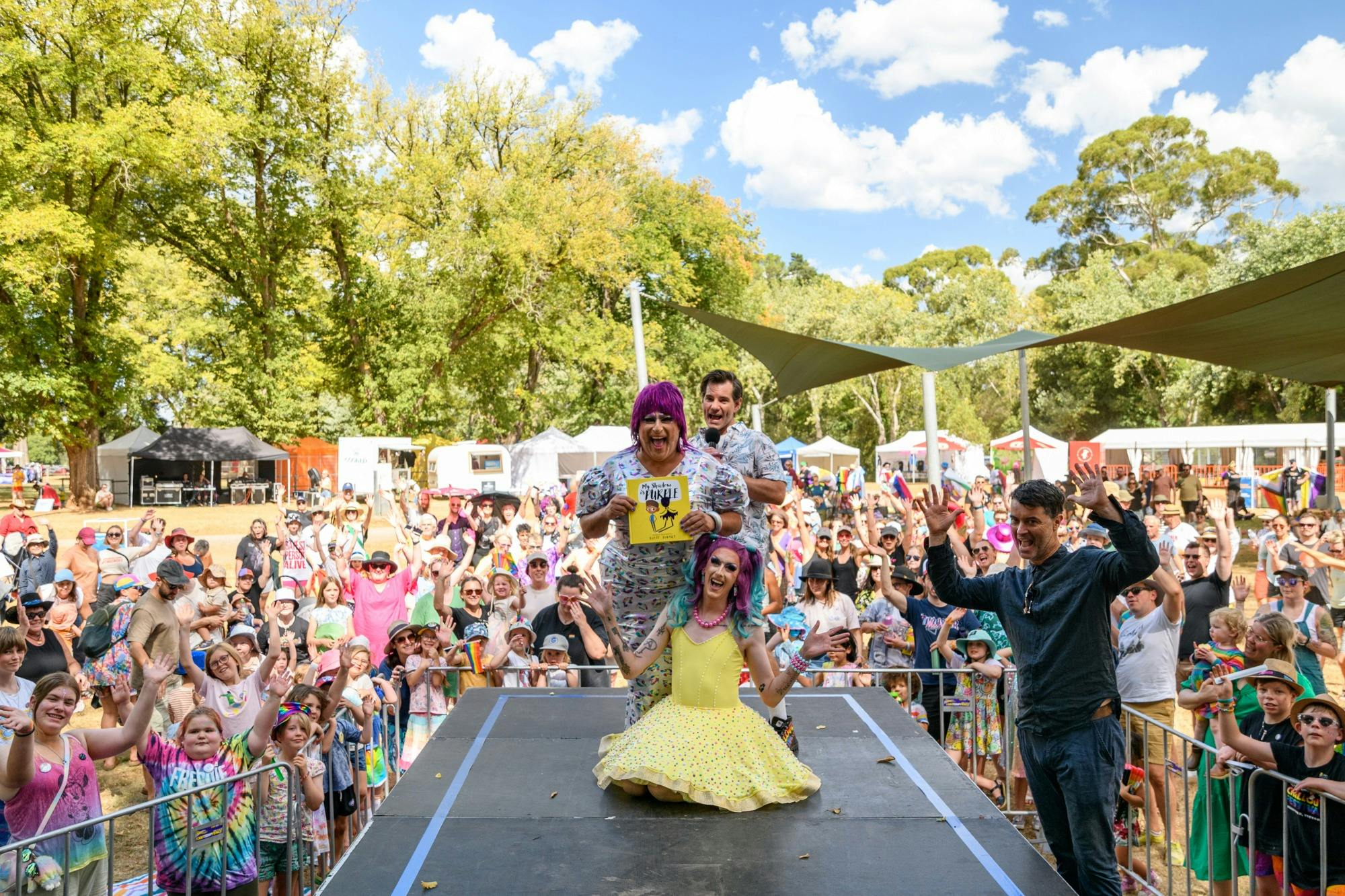 Two drag queens and Nate Byrne stand on stage surrounded by rainbow families