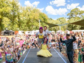 Two drag queens and Nate Byrne stand on stage surrounded by rainbow families