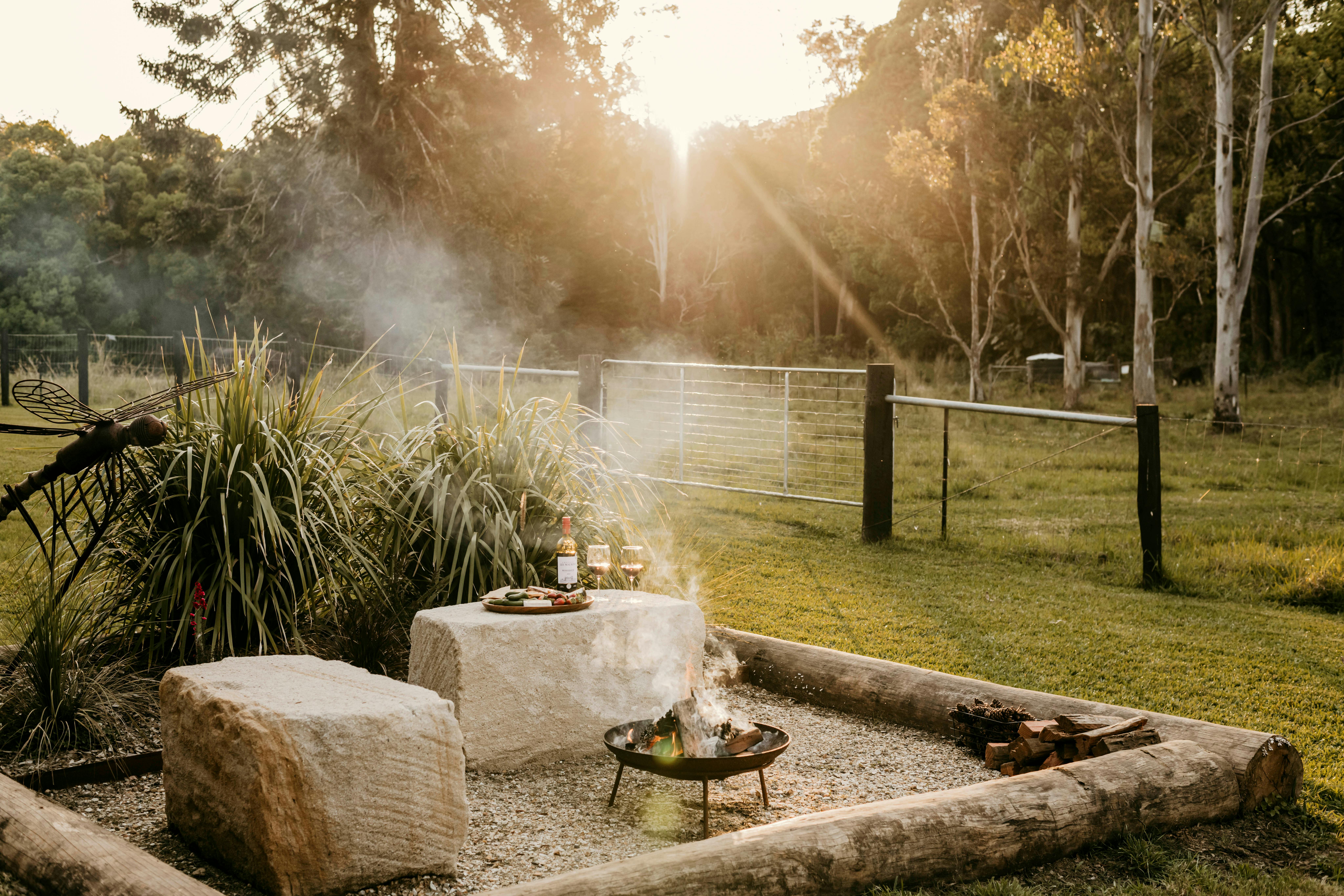 Firepit area with stone seating, wine, and sunset view at Contained in Nimbin’s grassy paddock.