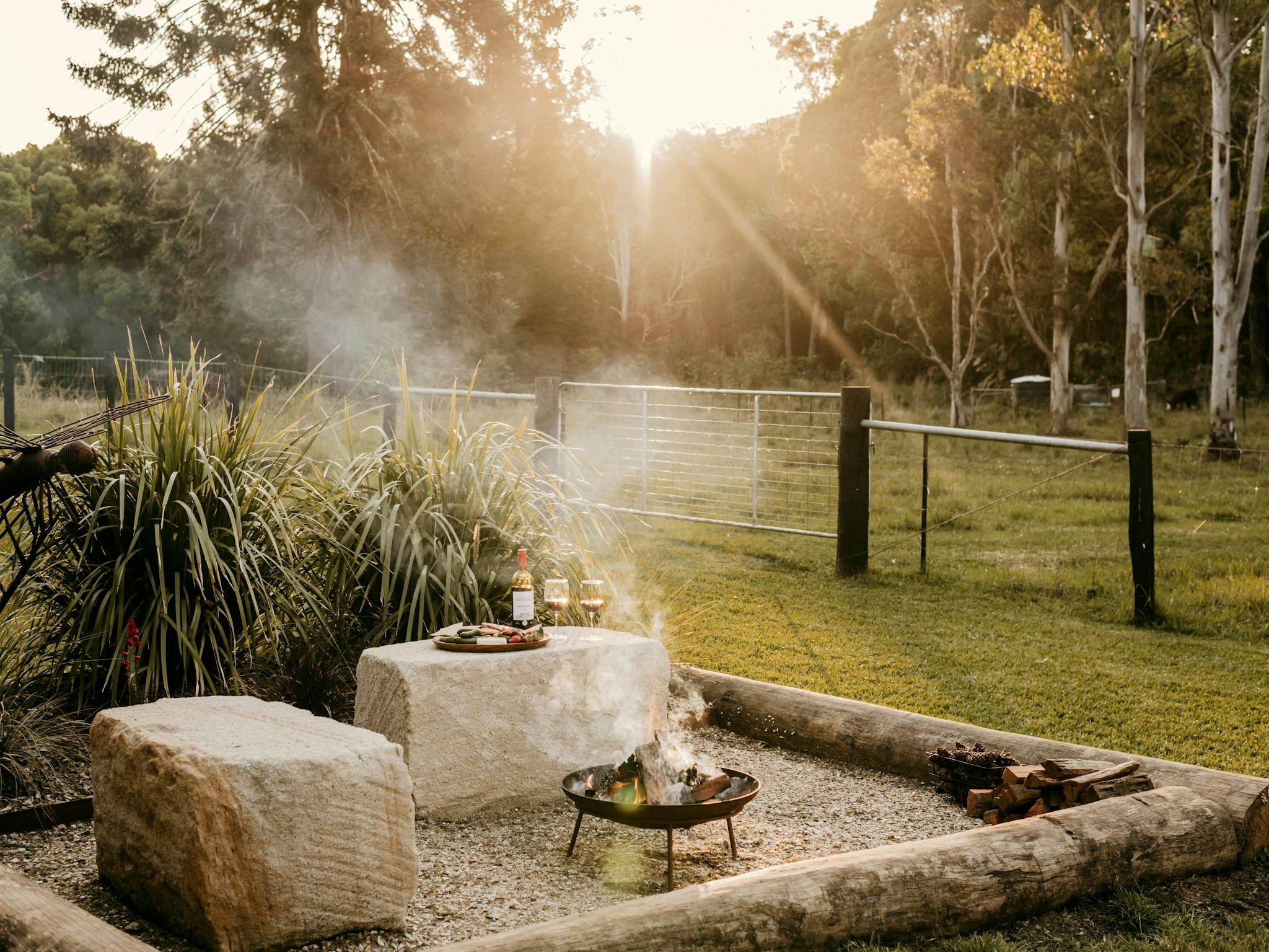 Firepit area with stone seating, wine, and sunset view at Contained in Nimbin’s grassy paddock.