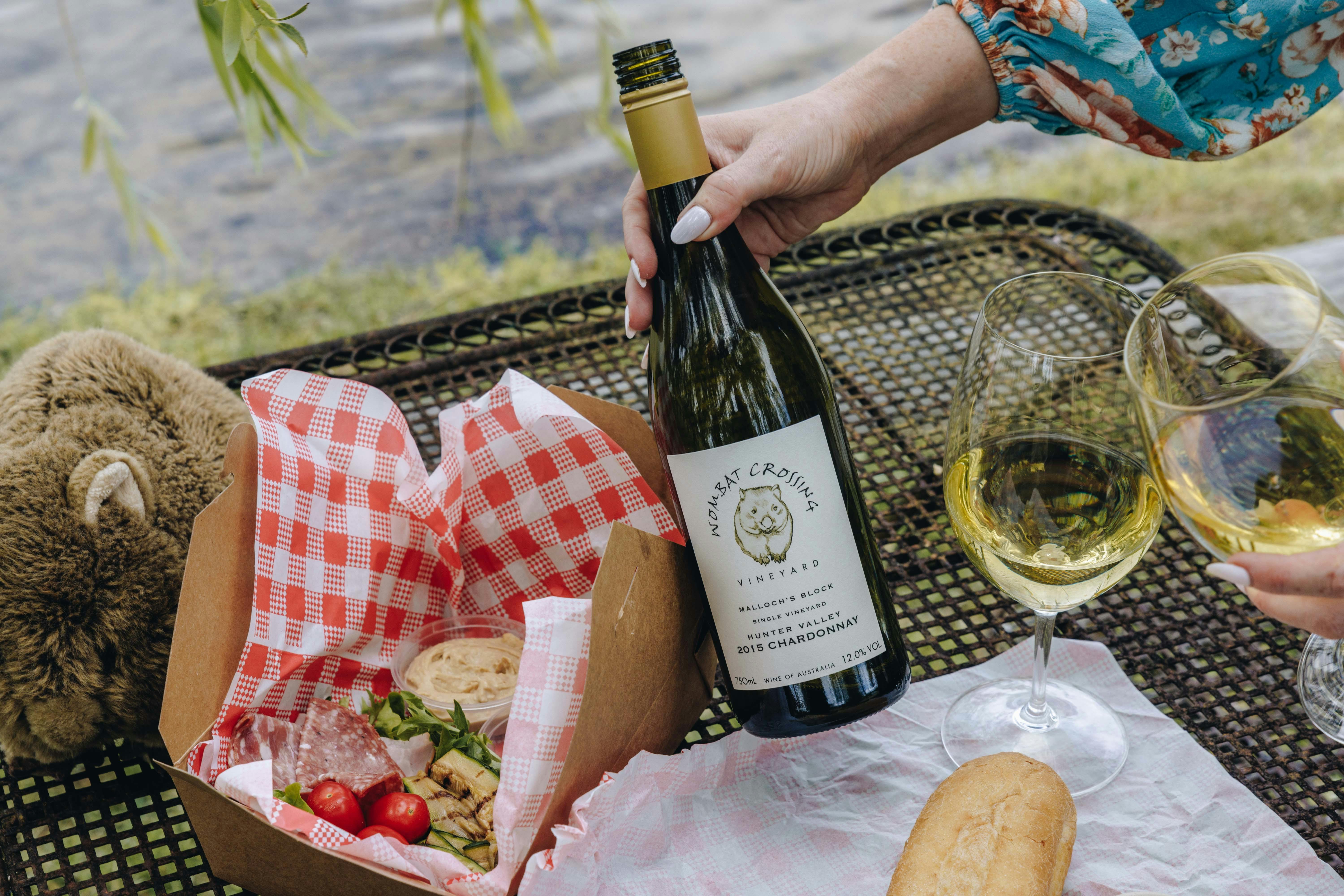A lady holds a wine bottle in her hand.  A gourmet picnic is laid out by a dam