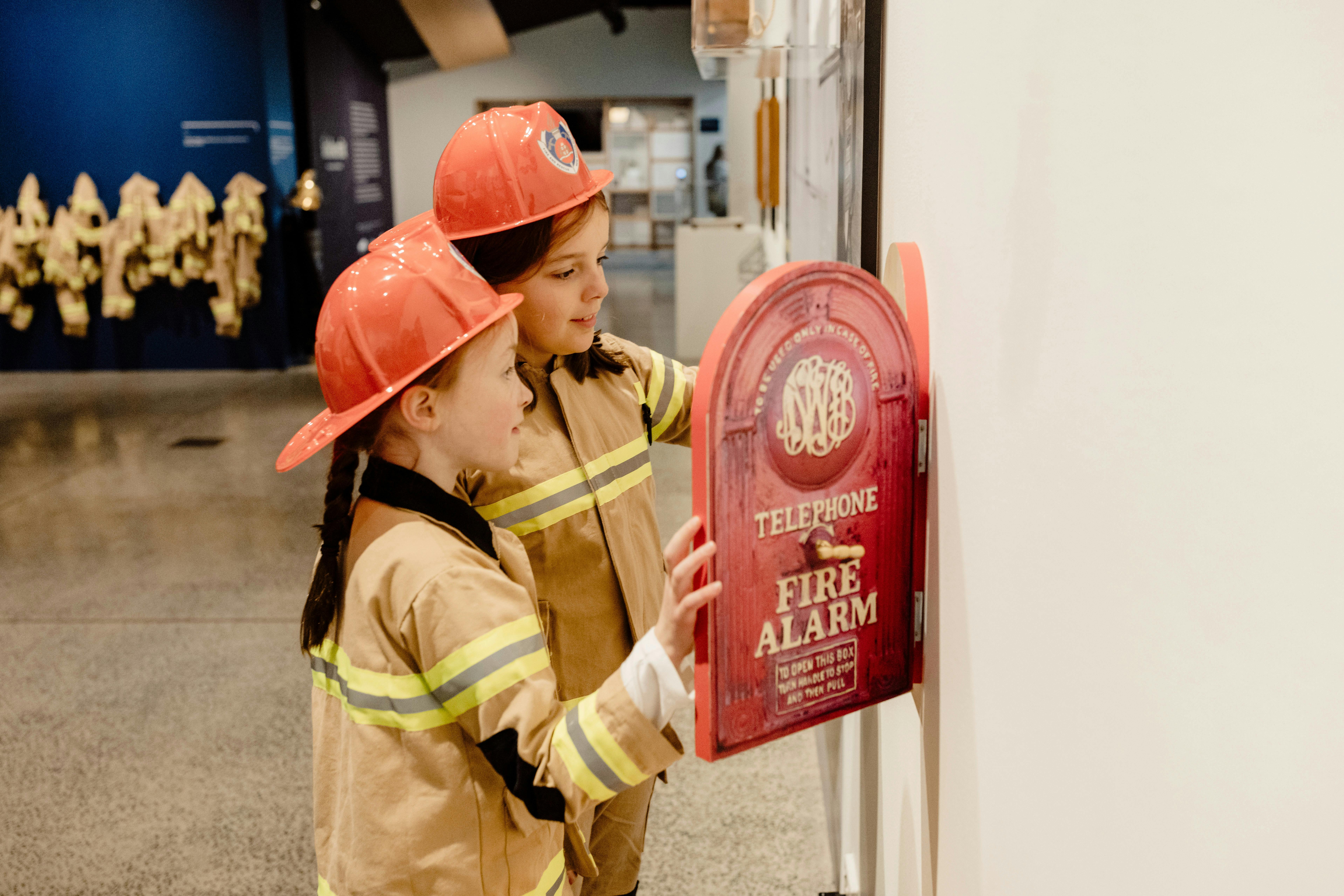 Two children dressed in fire fighter uniforms and hats looking at a replica fire alarm telephone